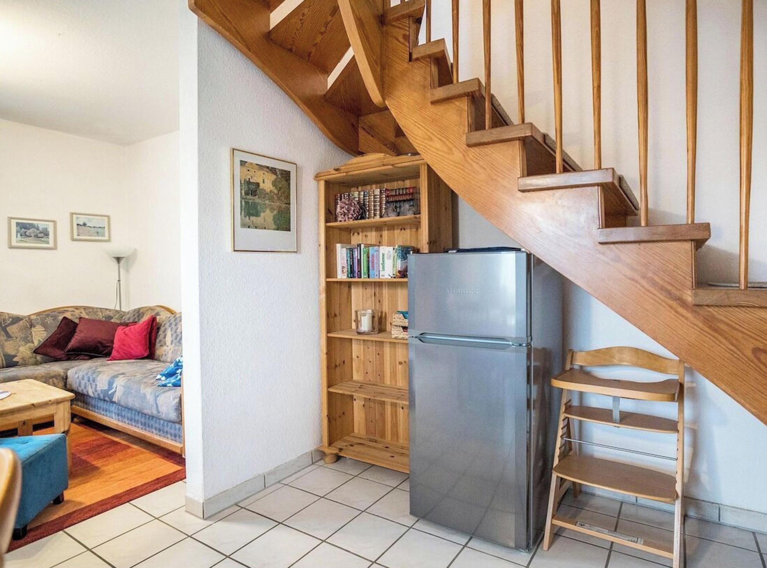 Een woonkamer met een houten trap, een bank, een bijzettafeltje, een boekenkast en een koelkast.
