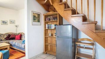 Een woonkamer met een houten trap, een bank, een bijzettafeltje, een boekenkast en een koelkast.