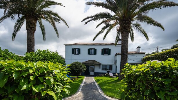 A white building with a tiled roof, a paved pathway leading to the entrance, and lush greenery on either side. - Vrbo Property (Rabo de Peixe)