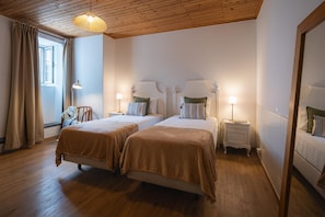 Une chambre d’hôtel avec deux lits, un plancher en bois, un plafond avec des poutres apparentes et une grande fenêtre avec des rideaux.