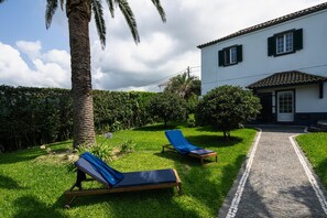 A property with a paved pathway leading to a white building with green shutters, two lounge chairs on a lawn, and a palm tree.