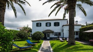 A two-story house with a paved pathway leading to the entrance, surrounded by lush greenery and palm trees. - Vrbo Property (Rabo de Peixe)