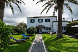 Une maison à deux étages, avec un chemin pavé menant à l’entrée, entourée d’une végétation luxuriante et de palmiers.