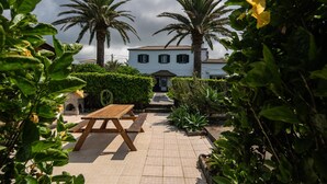 A courtyard with a wooden table, palm trees, and a building with a tiled roof. - Vrbo Property (Rabo de Peixe)