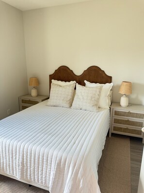 Een net opgemaakt bed met een gewatteerde witte hoes, twee bijpassende kussens en een houten hoofdbord. Aan beide zijden van het bed staan nachtkastjes met lampen.