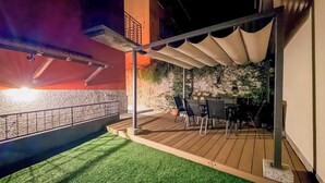 Un espace extérieur aménagé en terrasse, avec un sol en bois, du gazon synthétique, une table entourée de chaises et un mur en pierre en arrière-plan.