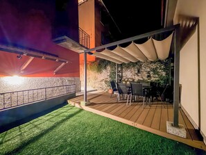 Un área de patio exterior con cubierta de madera, césped artificial, una mesa con sillas y un muro de piedra de fondo.