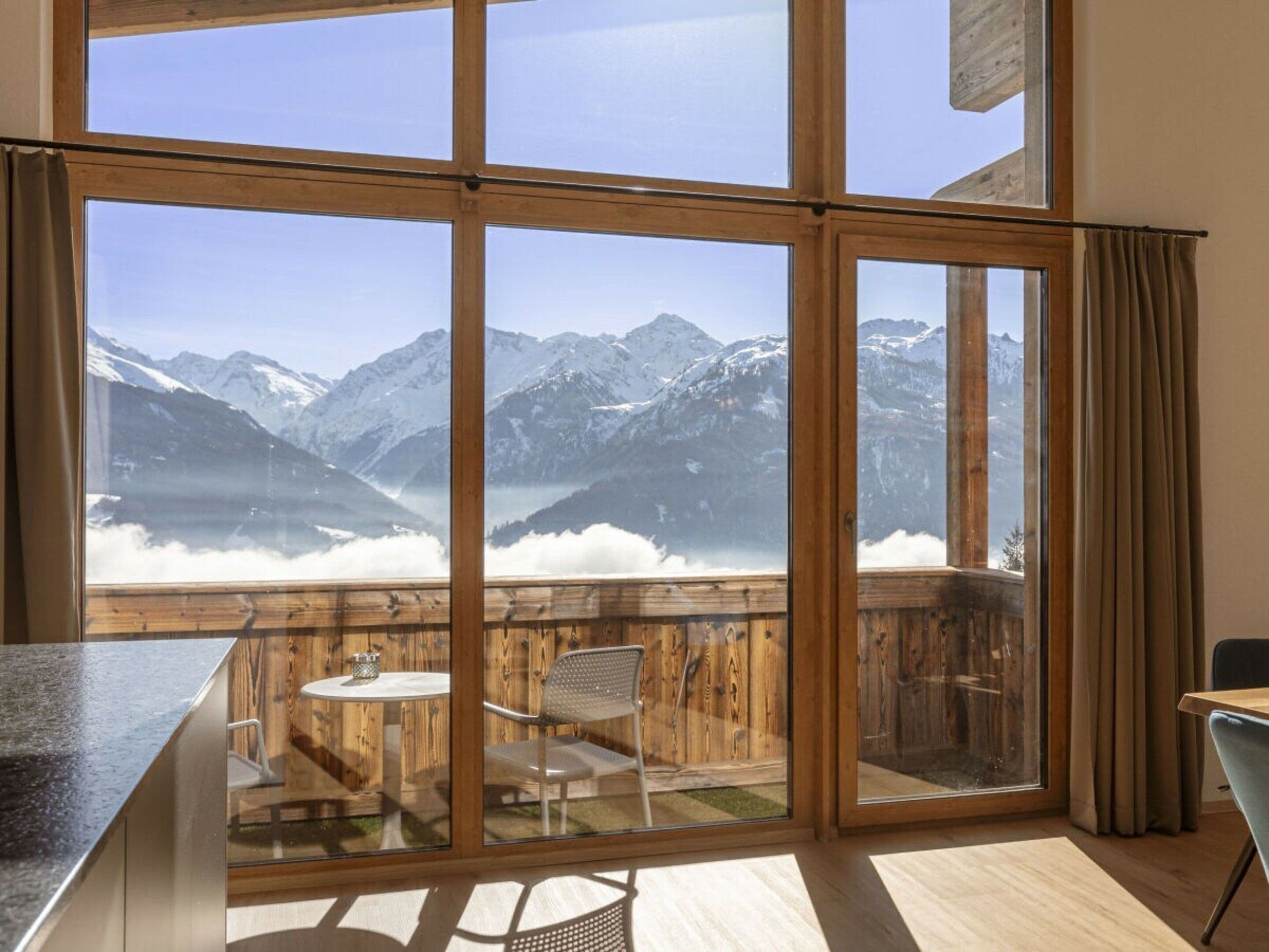 Ein Zimmer mit großen Fenstern und Blick auf die Berge, eine Holzdiele mit Tisch und Stühlen sowie eine moderne Küchenzeile.