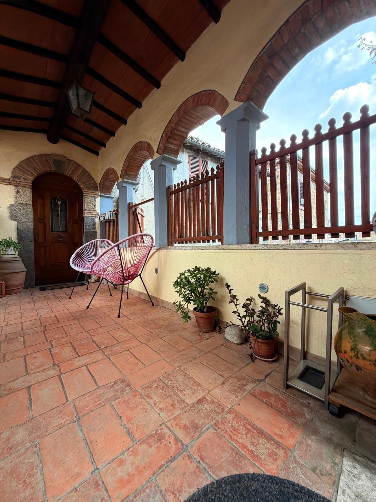 Un patio cubierto con baldosas de terracota, una puerta de madera, una silla rosa y plantas en macetas.
