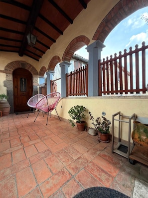 A covered patio with terracotta tiles, a wooden door, a pink chair, and potted plants.
