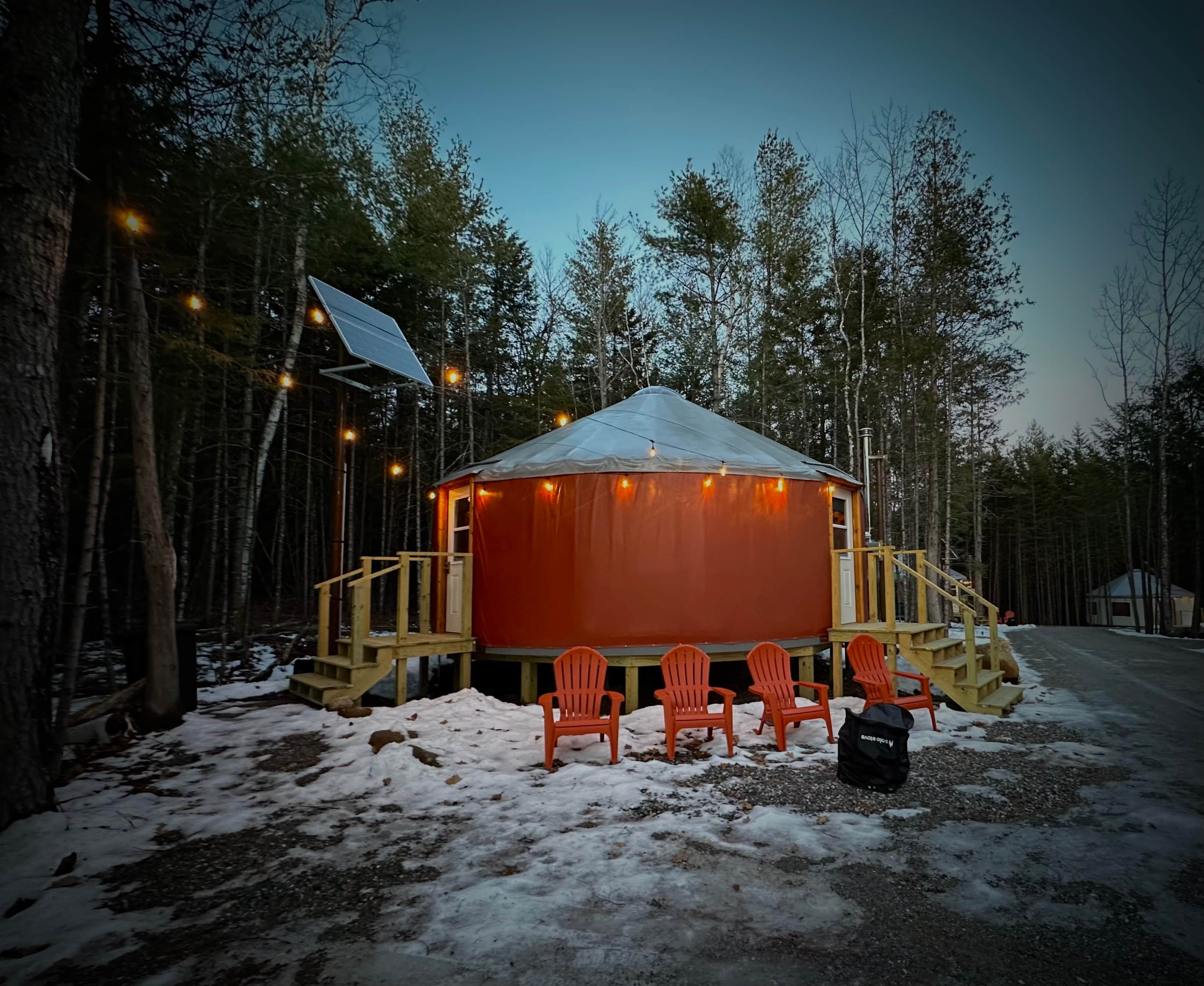 A wooden cabin with a snow-covered ground, surrounded by trees, and equipped with outdoor seating and a solar panel.