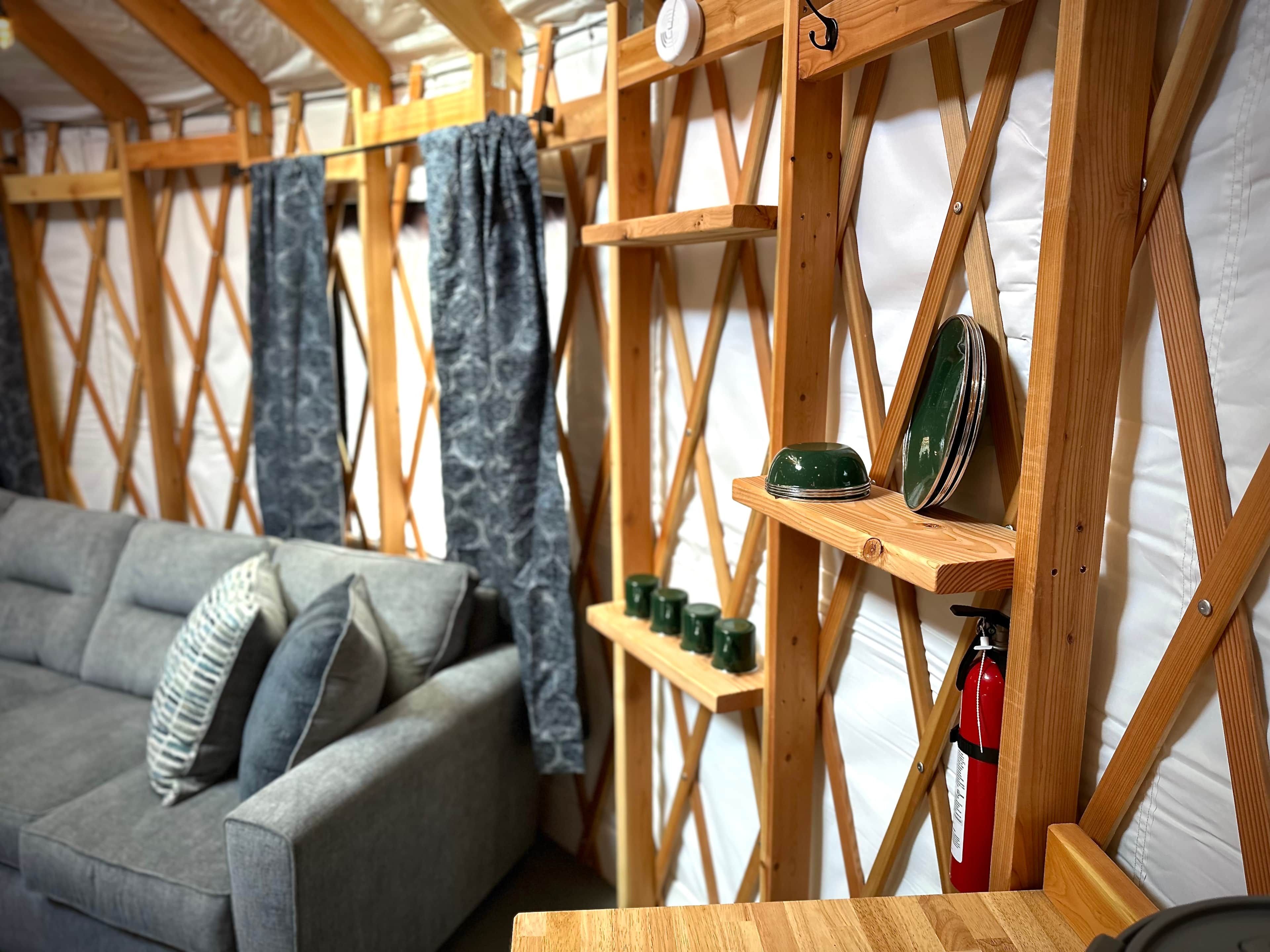 Ein gemütliches Zimmer mit sichtbarer Holzbalkendecke, einem grauen Sofa und Regalen mit grünen Geschirrgegenständen sowie einem Feuerlöscher.