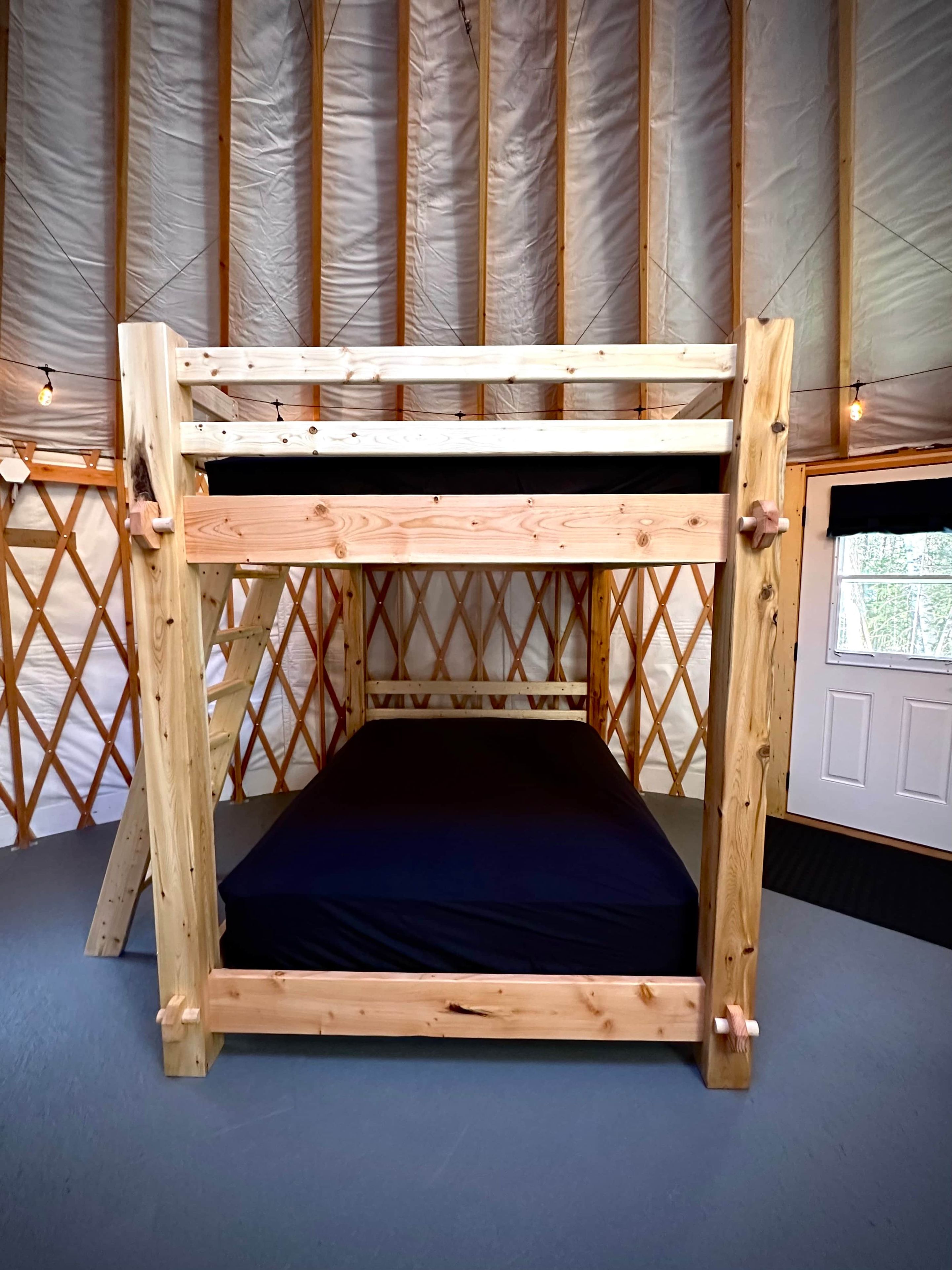 A bunk bed with a wooden frame and a black mattress, set against a backdrop of a tent-like structure with a lattice design.