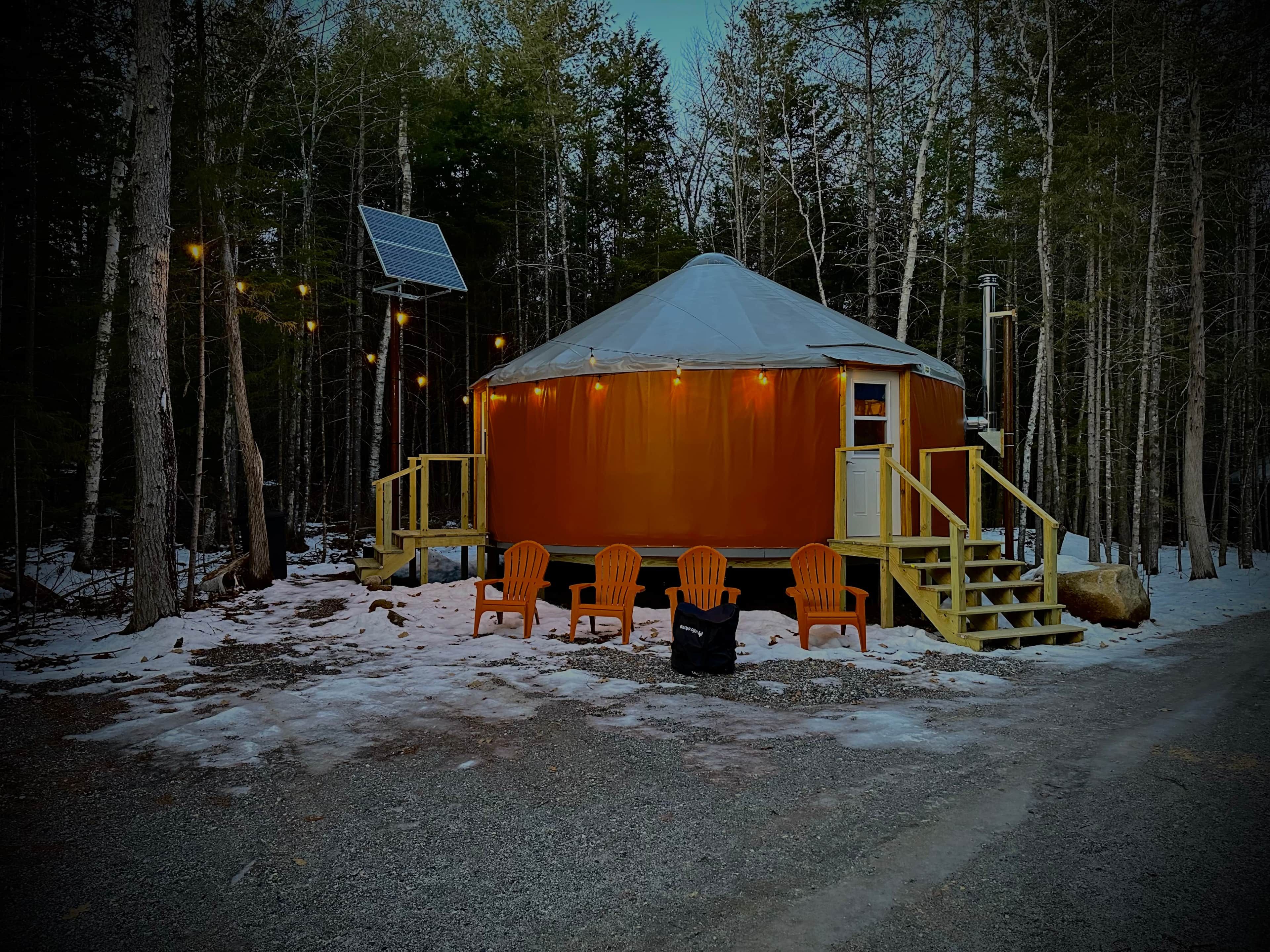En stuga med ett snötäckt tak, omgiven av en skog. Det finns en trappa av trä som leder upp till ingången, och en solpanel syns på taket.