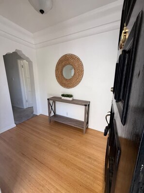 Habitació amb sòl de fusta, un mirall rodó a la paret, una taula petita amb una planta i una porta amb marc negre.