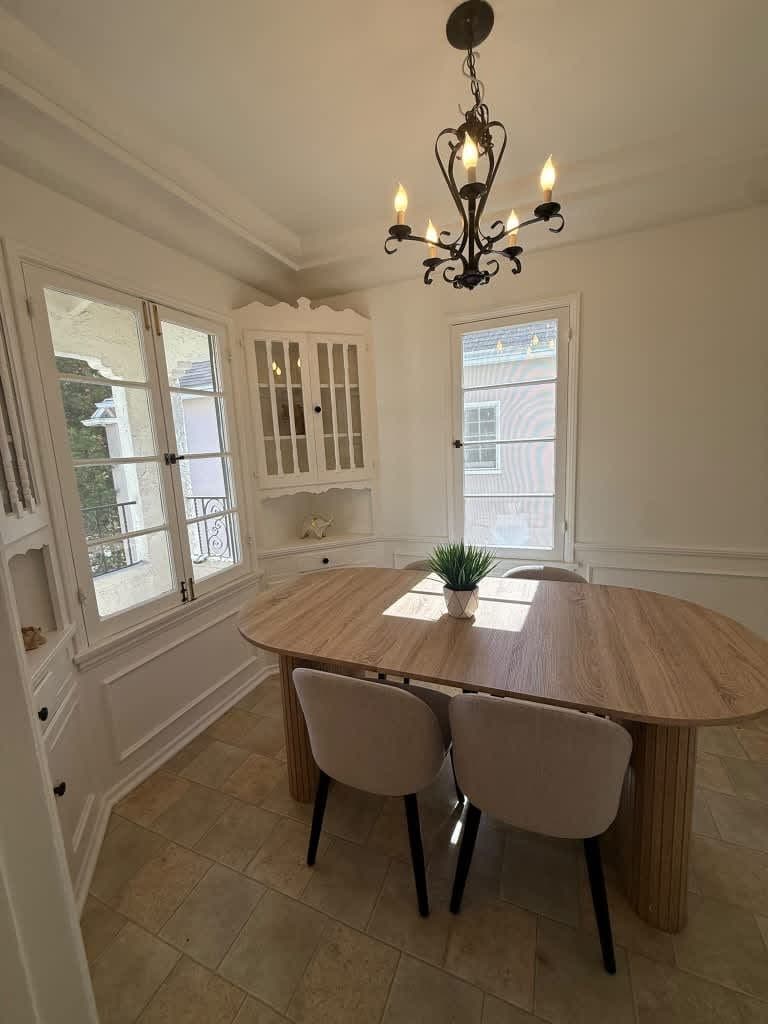 Uma sala de jantar com uma mesa redonda, duas cadeiras, um lustre e janelas amplas.