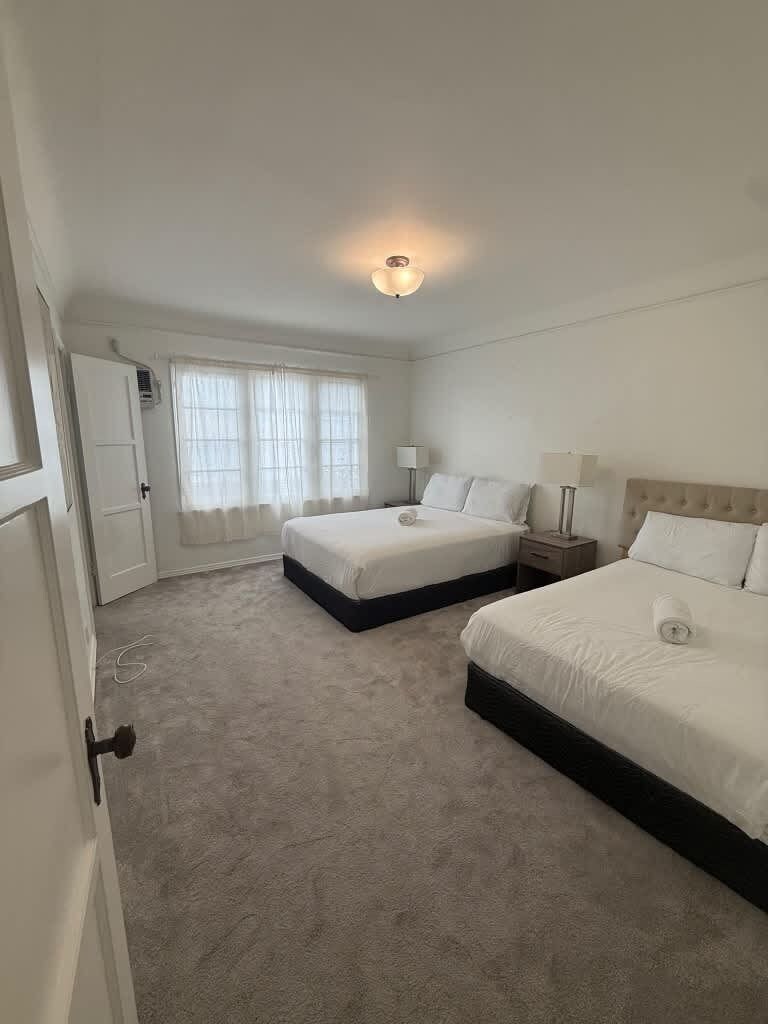 Quarto de hotel com duas camas, uma porta, uma janela com cortinas e uma luminária no teto.