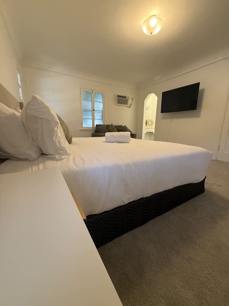 A bedroom with a bed, a bedside table, a flat-screen TV mounted on the wall, and a window with blinds.