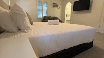 A bedroom with a bed, a bedside table, a flat-screen TV mounted on the wall, and a window with blinds.
