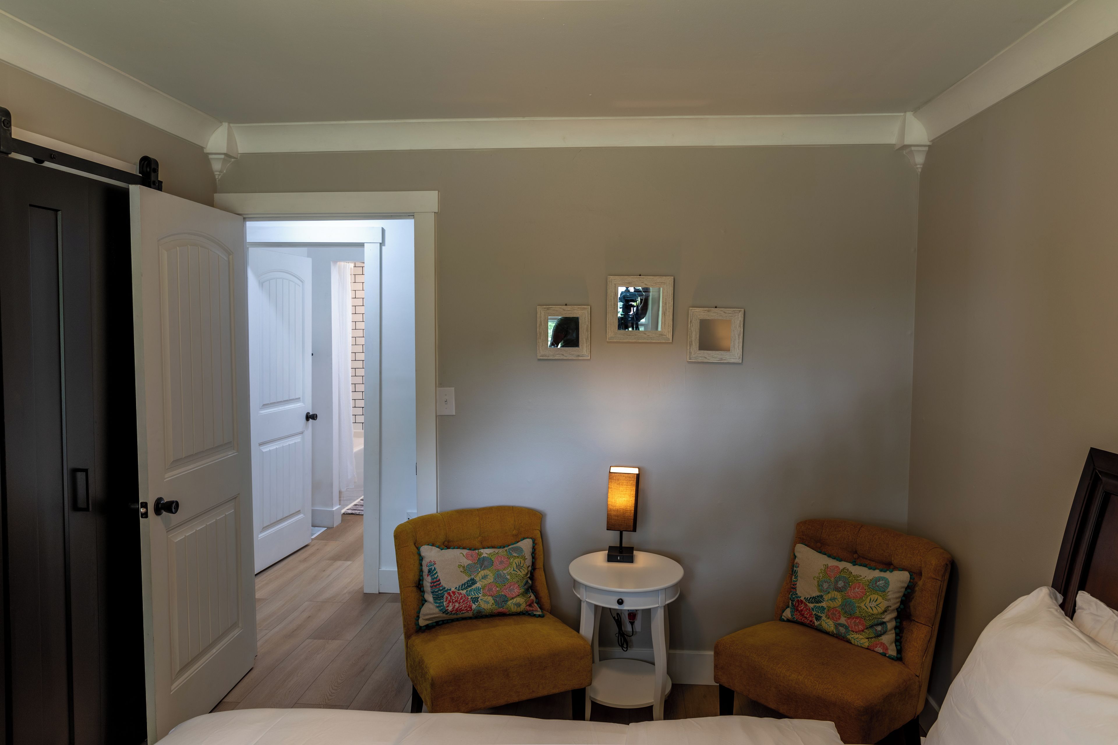 Quarto de hotel com uma cama, duas poltronas, uma mesinha de centro com abajur e uma porta que dá para outro quarto.