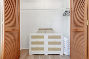 A white dresser with six drawers and a wicker front panel, positioned in a closet with wooden louvered doors. - Charming 3-bedroom house with heated pool and within 5 miles from the beach. (Vero Beach)