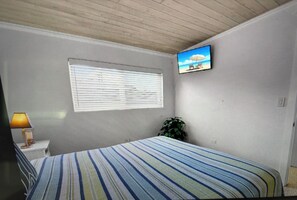 Une chambre à coucher avec un lit, une fenêtre avec des stores, un téléviseur fixé au mur et une lampe de chevet.