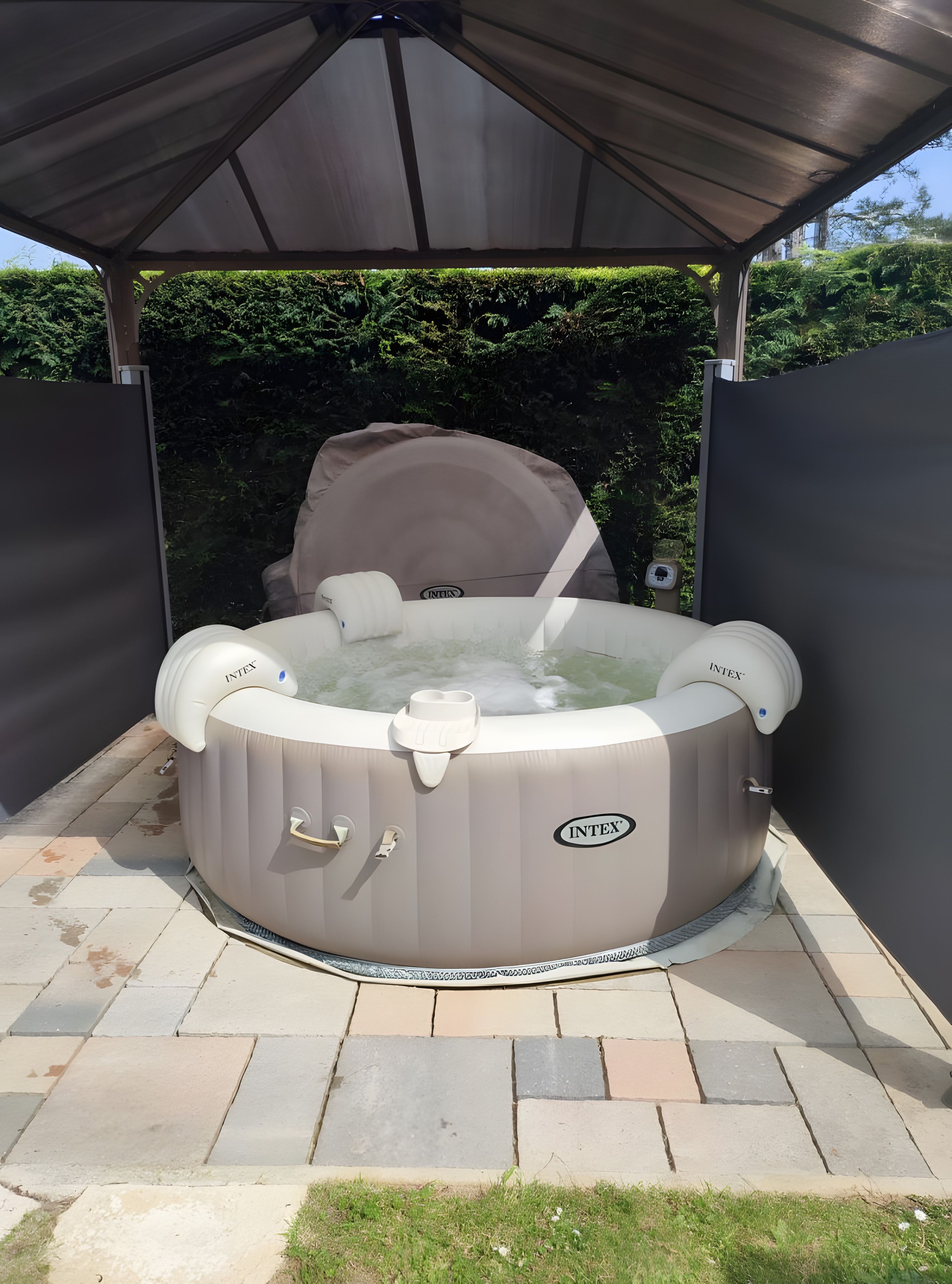 A hot tub under a covered patio with a stone-tiled floor and a hedge in the background.