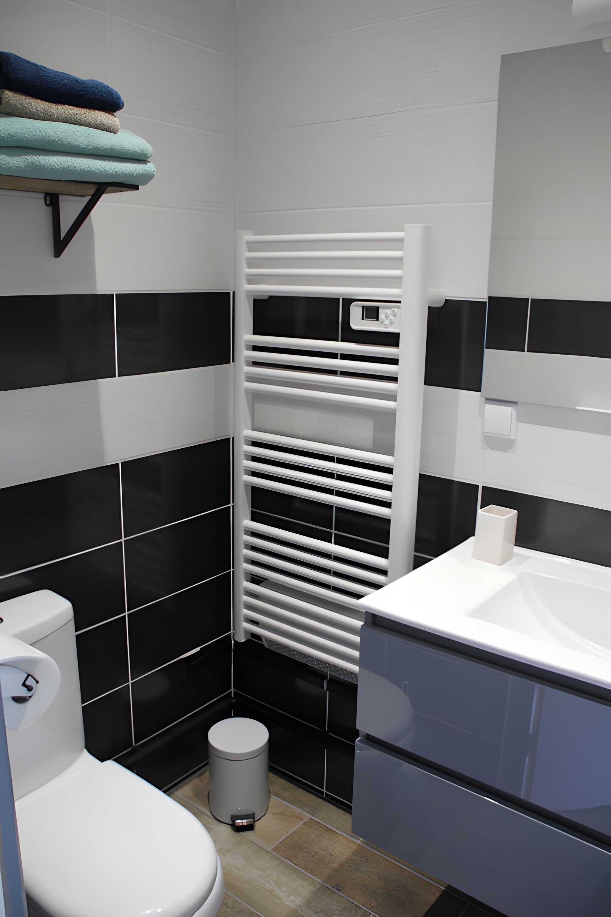 Ein Badezimmer mit einem weißen Waschbecken, schwarz-weißen Fliesen und einer weißen Toilette. Ein Handtuchhalter mit gefalteten Handtüchern ist ebenfalls vorhanden.