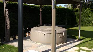 A covered outdoor seating area with a hot tub, surrounded by a garden with a pergola structure.
