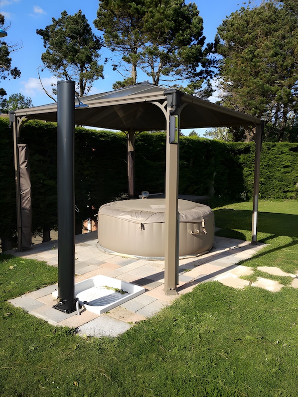 A covered outdoor seating area with a hot tub, surrounded by a garden with a pergola structure.