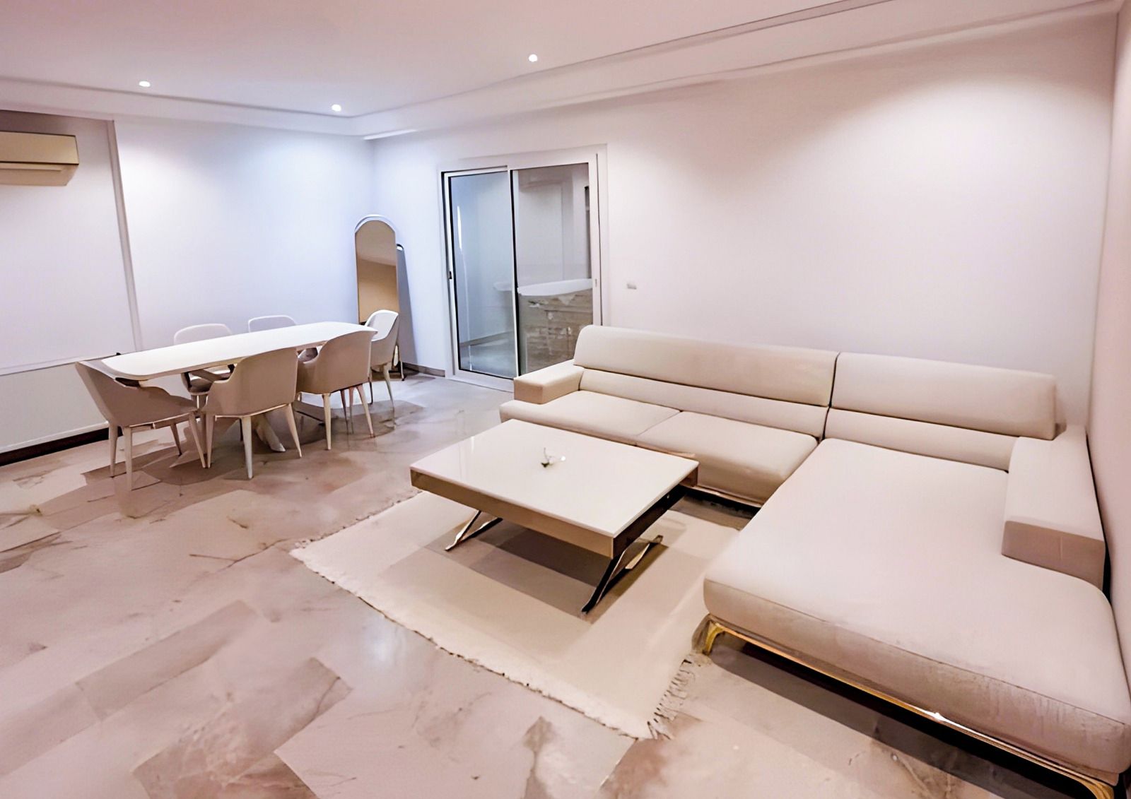 A modern living room with a sofa, a coffee table, a dining table with chairs, and a sliding glass door leading to an outdoor area.