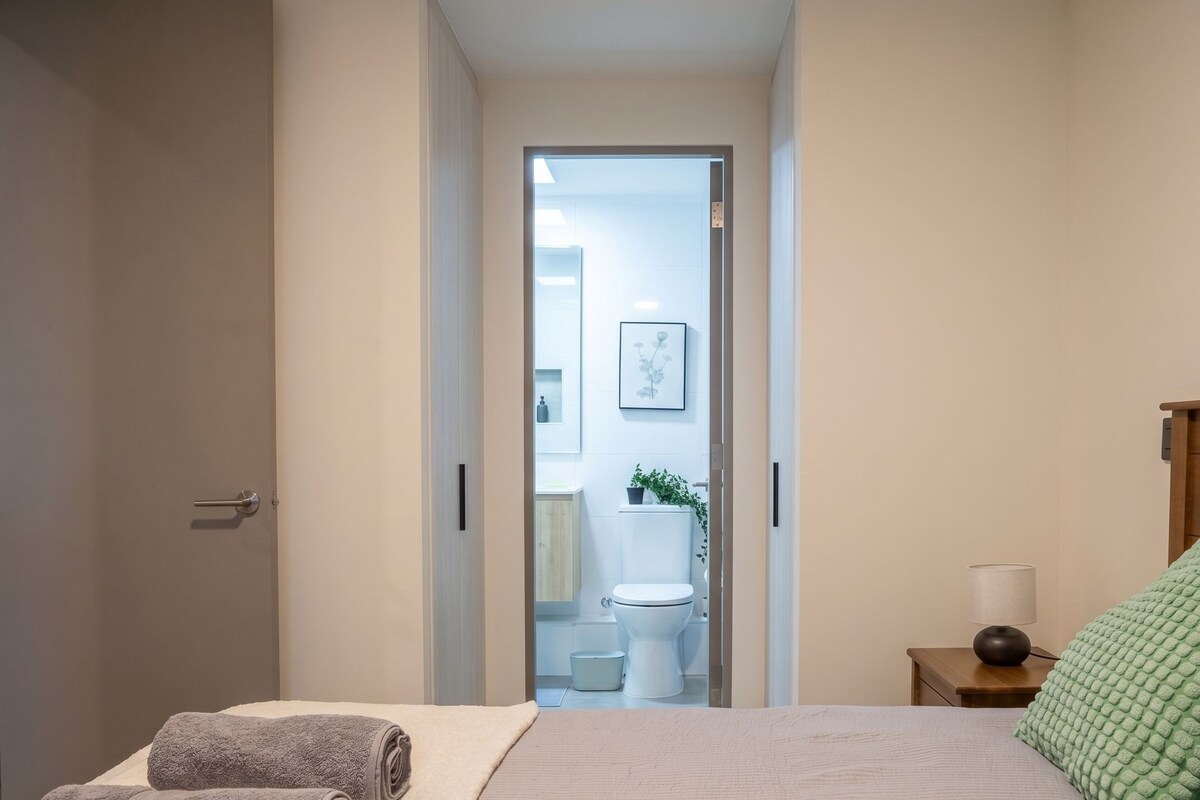 A bedroom with a bed, two rolled towels, a bedside table with a lamp, and a view into the bathroom.