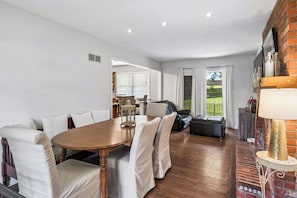 Uma sala de jantar com mesa de madeira e cadeiras estofadas brancas, lareira de tijolos e vista para um jardim através de uma porta de vidro.