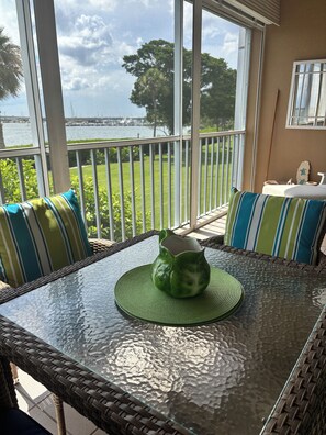 Ein Tisch mit Glasplatte, eine grüne Vase, Korbstühle mit gestreiften Kissen und Blick auf ein Gewässer durch große Fenster.
