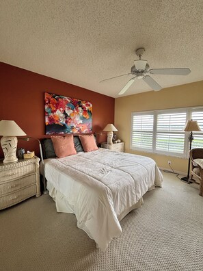 Ein Schlafzimmer mit Bett, Deckenventilator, Fenster mit Jalousien und einem Bild an der Wand.