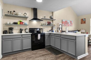 A kitchen with grey cabinets, a black stove, and a white countertop. There are shelves above the counter holding various kitchen items.