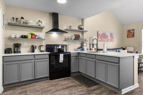 A kitchen with grey cabinets, a black stove, and a white countertop. There are shelves above the counter holding various kitchen items.