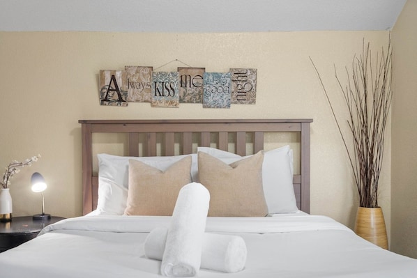 A neatly made bed with white linens and beige pillows, a bedside lamp, and a decorative wall piece above the bed.