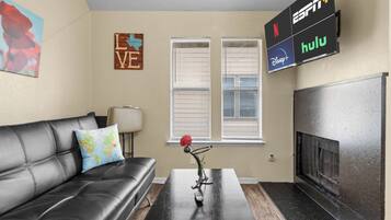 Uma sala de estar com um sofá de couro preto, uma mesa de centro, uma TV fixada na parede exibindo serviços de streaming e uma lareira.