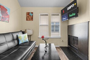 A living room with a black leather sofa, a coffee table, a wall-mounted TV displaying streaming services, and a fireplace.