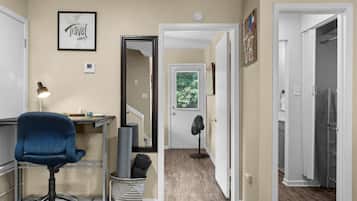 Corredor com escrivaninha, cadeira e espelho. Há uma porta que dá para outro quarto com janela.