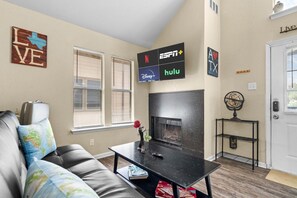 A living room with a fireplace, a couch, a coffee table, and a TV mounted on the wall displaying ESPN, Disney+, and Hulu logos.