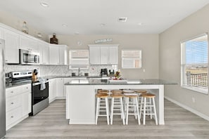A modern kitchen with white cabinetry, a central island with stools, a microwave, and a stove. - Blue Lobster Lodge | Beach Hair, Zero Cares (Bolivar Peninsula)
