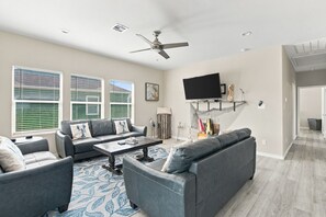 A modern living room with a ceiling fan, a flat-screen TV mounted on a wall, and a fireplace with a mantel. - Blue Lobster Lodge | Beach Hair, Zero Cares (Bolivar Peninsula)