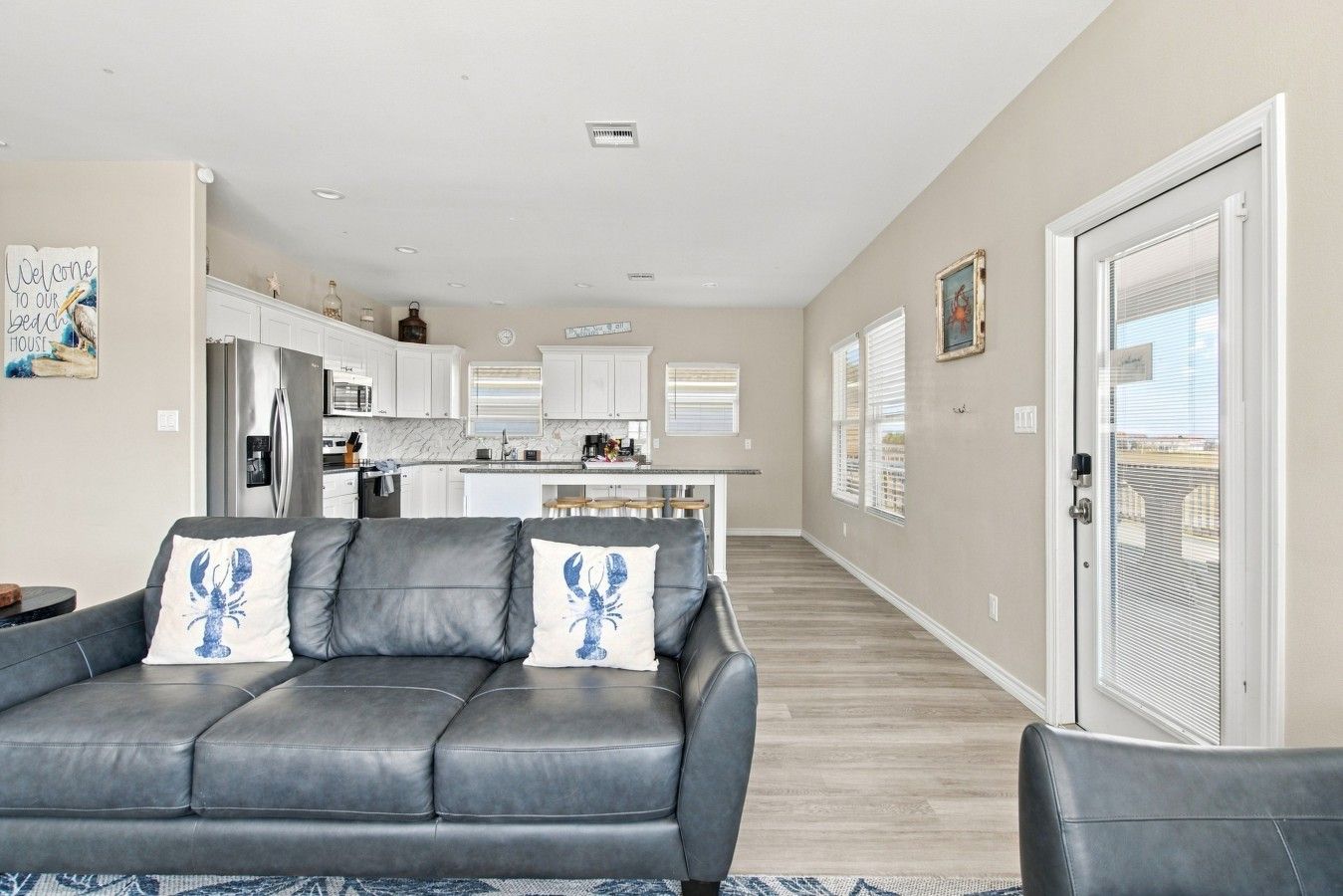 A modern living room with a grey leather sofa, a kitchen in the background, and a sliding glass door leading to a balcony.