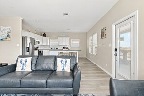 A modern living room with a grey leather sofa, a kitchen in the background, and a sliding glass door leading to a balcony. - Blue Lobster Lodge | Beach Hair, Zero Cares (Bolivar Peninsula)