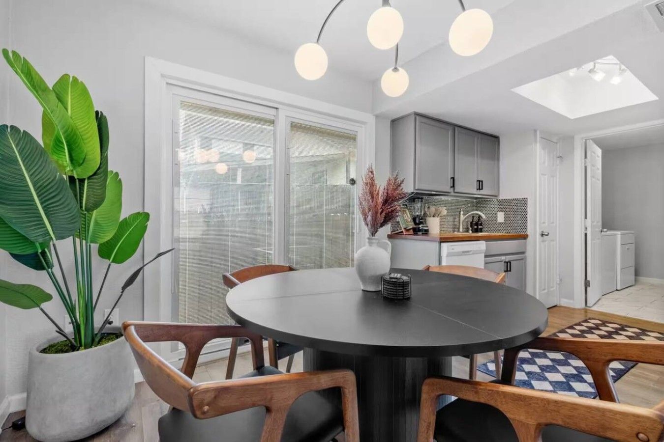 A modern kitchen with white cabinets, a dining area with a round table and wooden chairs, and a large potted plant.