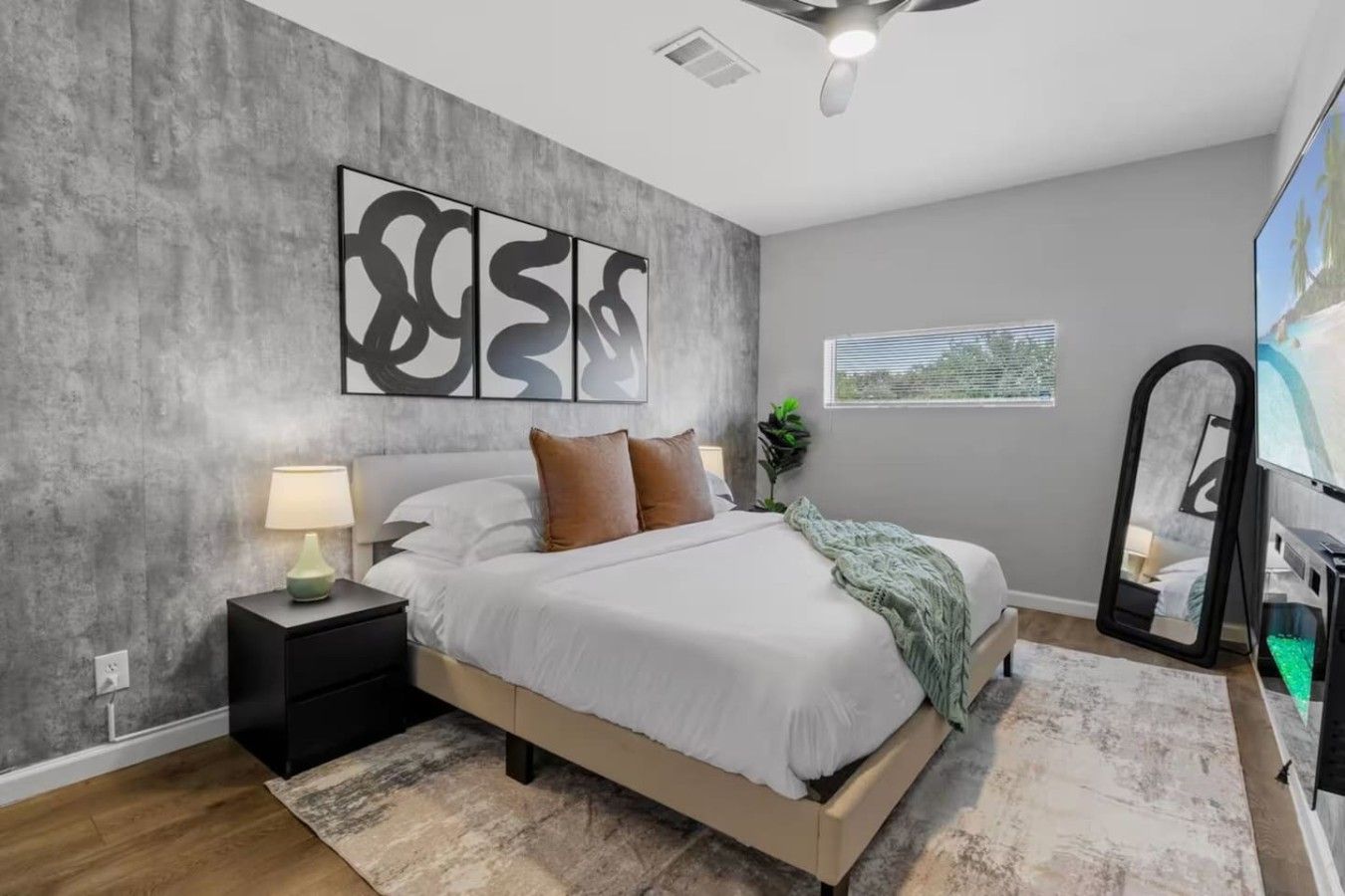A modern bedroom with a bed, bedside table, lamp, artwork, window, and a mirror.