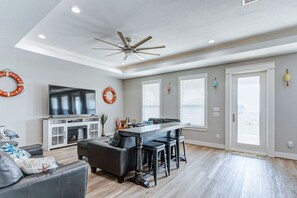 Un salon spacieux doté d’un grand téléviseur à écran plat, d’un canapé en cuir foncé, d’un coin repas avec un tabouret de bar et d’un ventilateur de plafond.