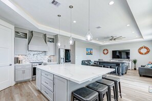 A modern kitchen with a large island, bar stools, and a dining area with a TV. - Royal Beach Retreat | Sleeps 16 | 4Bd/2.5Bth (Bolivar Peninsula)